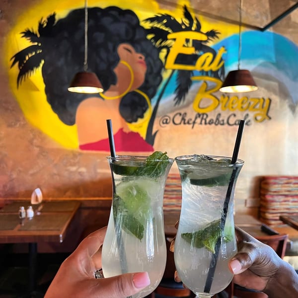 Two hands holding mojitos with lime and mint, against a vibrant mural at Chef Rob's Cafe.
