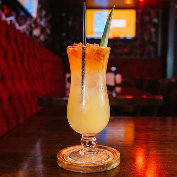 Tropical cocktail with pineapple leaf and straw on a wooden table in a dimly lit restaurant.