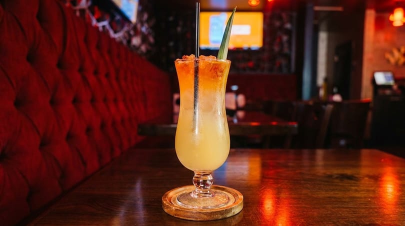 Tropical cocktail with pineapple leaf and straw on a wooden table in a dimly lit restaurant.