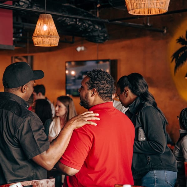 People socializing in a dimly lit restaurant with woven pendant lights and a palm tree mural.