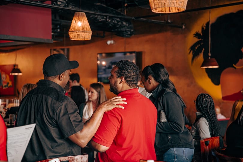 People socializing in a dimly lit restaurant with woven pendant lights and a palm tree mural.