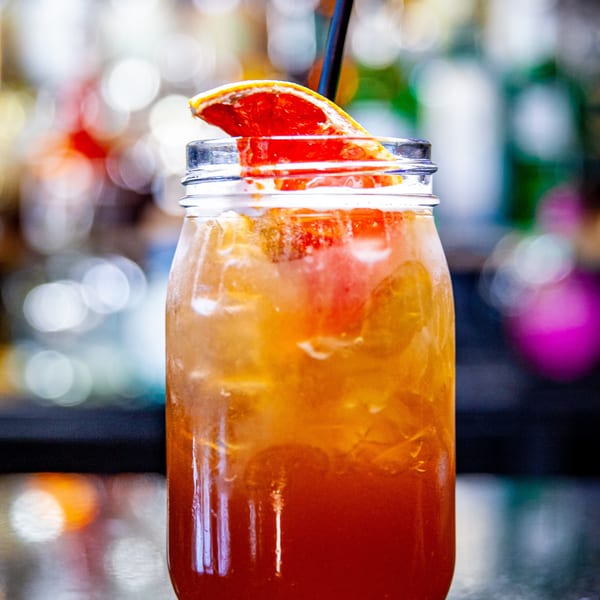 A vibrant orange and red cocktail in a mason jar with ice, a black straw, and a grapefruit slice.