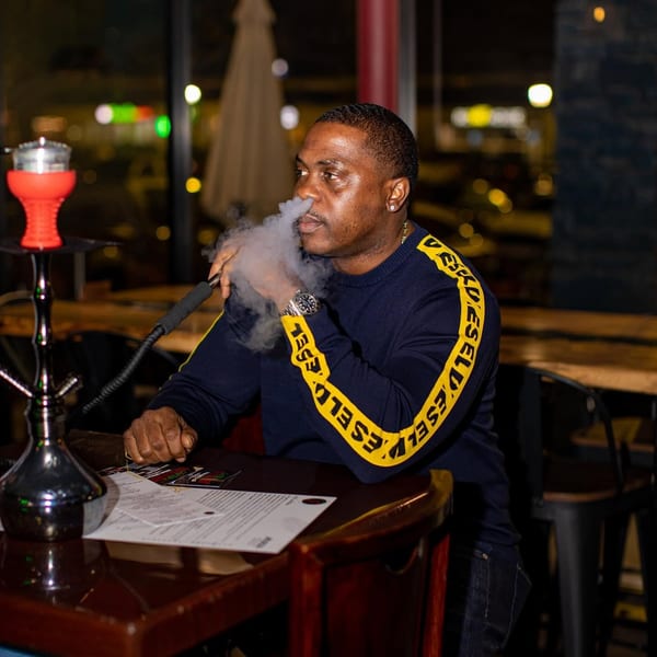 Man in a dark blue and yellow sweater exhaling hookah smoke at a high-top table.