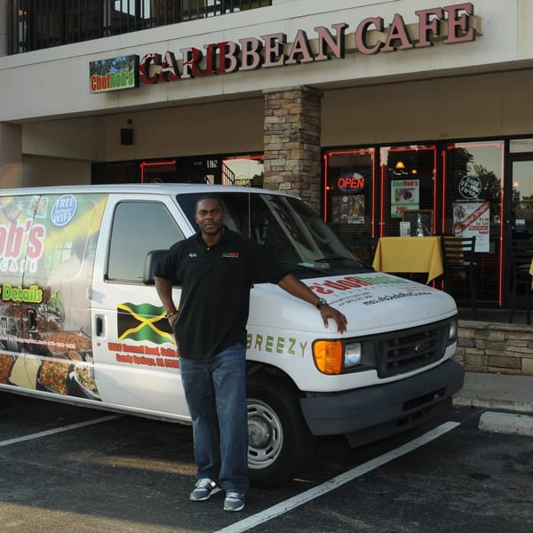 A man stands proudly beside a Chef Rob's Caribbean Cafe catering van outside the restaurant.