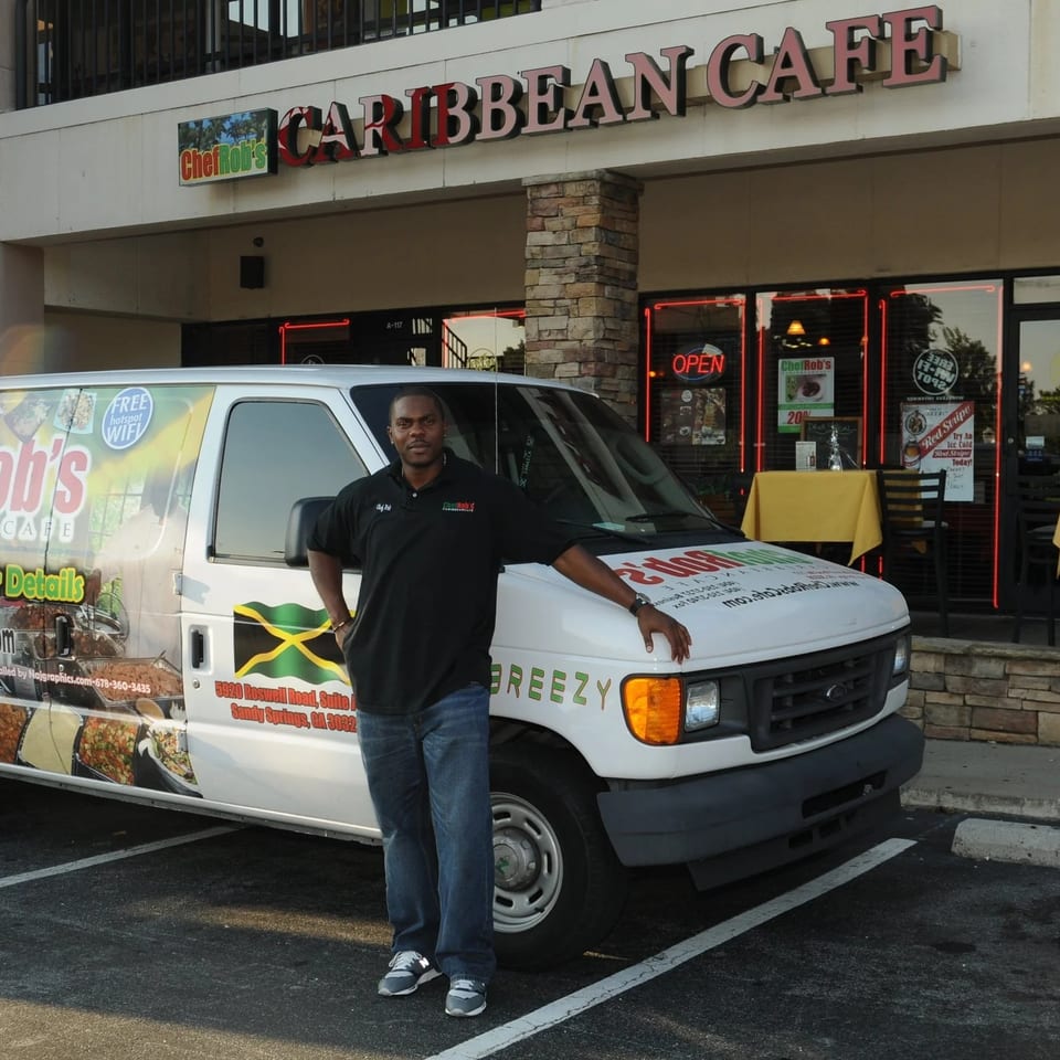 A man stands proudly beside a Chef Rob's Caribbean Cafe catering van outside the restaurant.