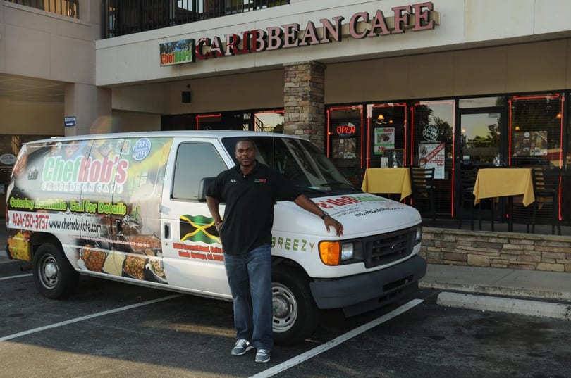 A man stands proudly beside a Chef Rob's Caribbean Cafe catering van outside the restaurant.