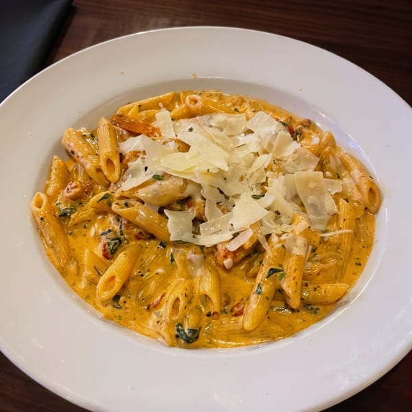 Penne pasta in a creamy orange sauce with herbs and shaved Parmesan cheese in a white bowl.