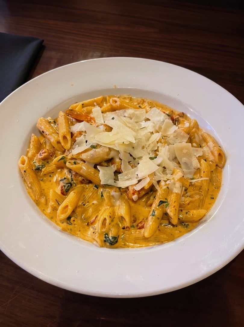 Penne pasta in a creamy orange sauce with herbs and shaved Parmesan cheese in a white bowl.