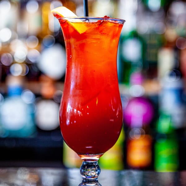 A vibrant red cocktail with a lemon slice and straw sits on a bar, with blurred bottles in the background.