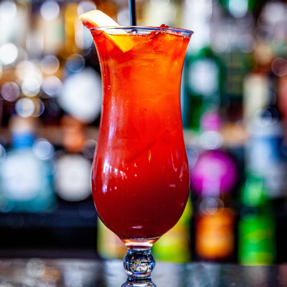 A vibrant red cocktail with a lemon slice and straw sits on a bar, with blurred bottles in the background.