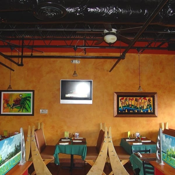 Restaurant interior with orange walls, exposed black ceiling, framed artwork, and set tables with green tablecloths.
