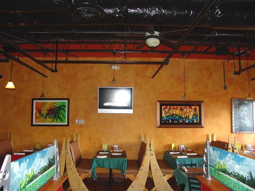 Restaurant interior with orange walls, exposed black ceiling, framed artwork, and set tables with green tablecloths.