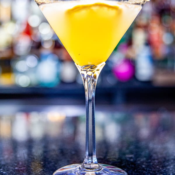 A vibrant yellow cocktail in a sugar-rimmed martini glass sits on a dark bar counter.