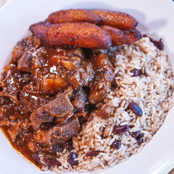 Oxtail stew with rice and peas, and fried plantains on a white plate.