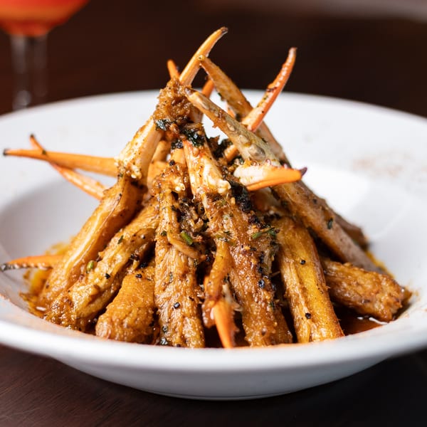A white bowl filled with seasoned crab legs, garnished with herbs and spices, with a drink in the background.