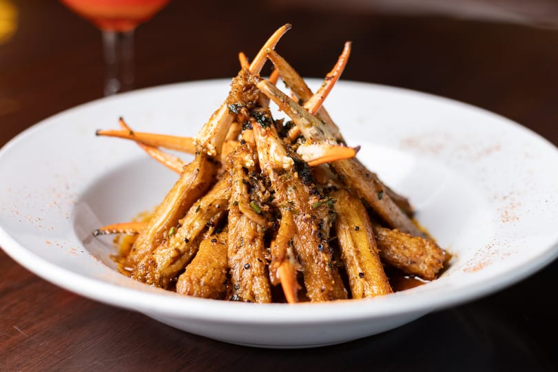 A white bowl filled with seasoned crab legs, garnished with herbs and spices, with a drink in the background.
