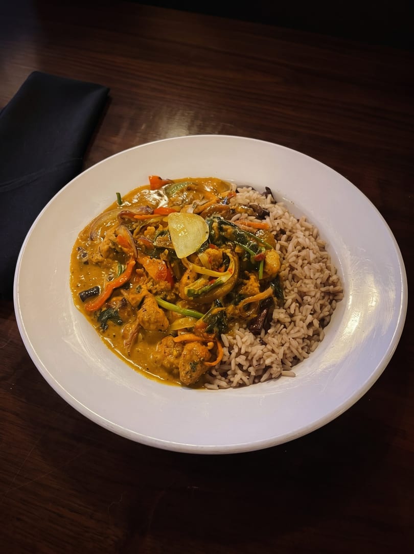 Curry chicken with vegetables and brown rice on a white plate, with a black napkin on a wooden table.