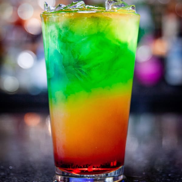 A vibrant, layered cocktail with green, yellow, and red liquids, topped with ice, on a dark bar.